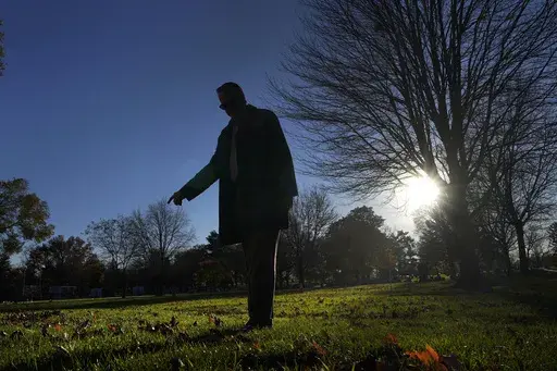 Cook County, Ill., Sheriff's Commander Jason Moran, who leads the sheriff's missing persons initiative stands in silhouette and points to a round cylinder that marks a grave of a person who self-identified as Seven, at the Mount Olivet Cemetery on Chicago's Far South Side Monday, Nov. 13, 2023. “That’s a horrible circumstance that someone could die and no one knows who they are. That’s why we pursue these cases so strongly, out of dignity,” says Moran, who oversees the sheriff’s missin