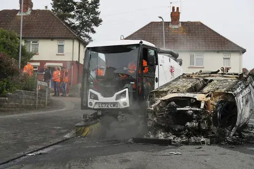 Council workers clear debris from the area immediately around a car that was set alight in Ely, Cardiff, Tuesday, May 23, 2023. Several dozen youths pelted police with objects and set cars ablaze on Monday evening in Cardiff in local unrest that erupted after two teenagers died in a road accident. (PA via AP)