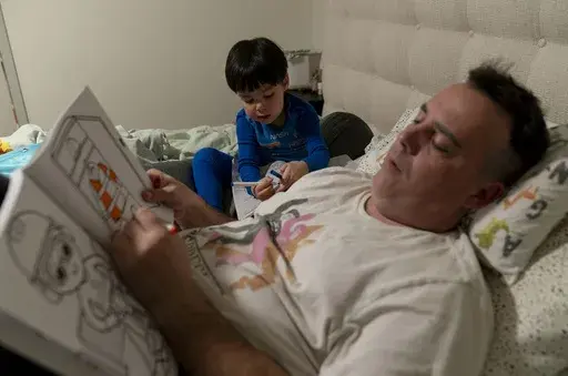 Scott Quinn and his 4-year-old son, Ethan, work on coloring books in a bed in Concord, Calif., Wednesday, Nov. 1, 2023. Ethan's parents are among those who have opted for a private daycare instead of a free “transitional kindergarten.” Quinn said he has been discouraged to see Ethan — one of the oldest kids in his daycare class — pick up the behavior of kids who are several years younger than him. “In retrospect, it would have been better to send him to school to be around kids his age