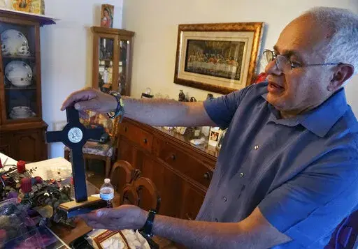 Bob Guerra, a Catholic deacon, holds a crucifix he got to commemorate the visit of Pope Francis to the U.S.-Mexican border at his house in El Paso, Texas, on Sunday, April 3, 2022. Guerra takes the crucifix alongside other liturgical items to help clergy celebrate Mass for hundreds of teens held at a shelter on the Army's Fort Bliss base after crossing the border without parents or guardians. (AP Photo/Giovanna Dell'Orto)