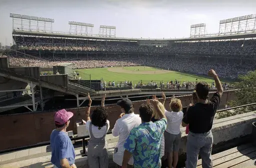 In this Monday, July 9, 1990, file photo, spectators watch an All-Star Game practice session from the roof of a building just outside Chicago's Wrigley Field. Booking hotels for baseball games during shoulder seasons like May, June or September can often be cheaper than the summer months. Additionally, save money by planning to attend weekday versus weekend games. (AP Photo/Seth Perlman, File)